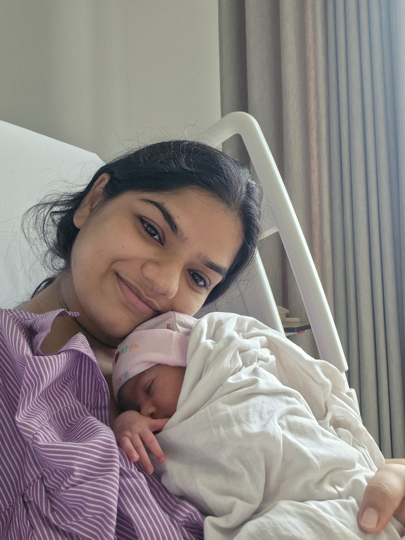 Shruti cuddling newborn Adira close against her in a hospital bed.