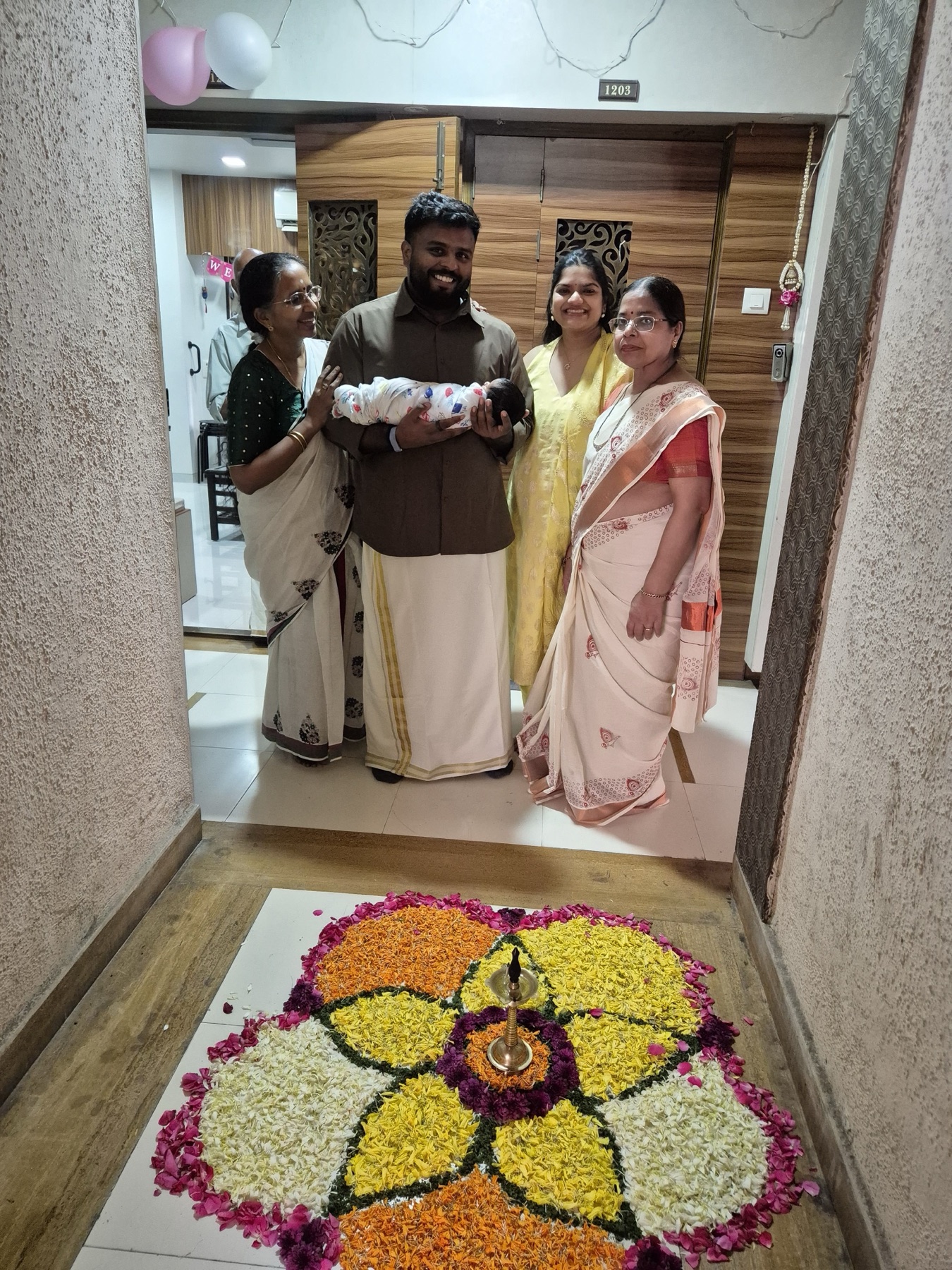 Family holding Adira at a decorated home doorway above a large flower pookalam.